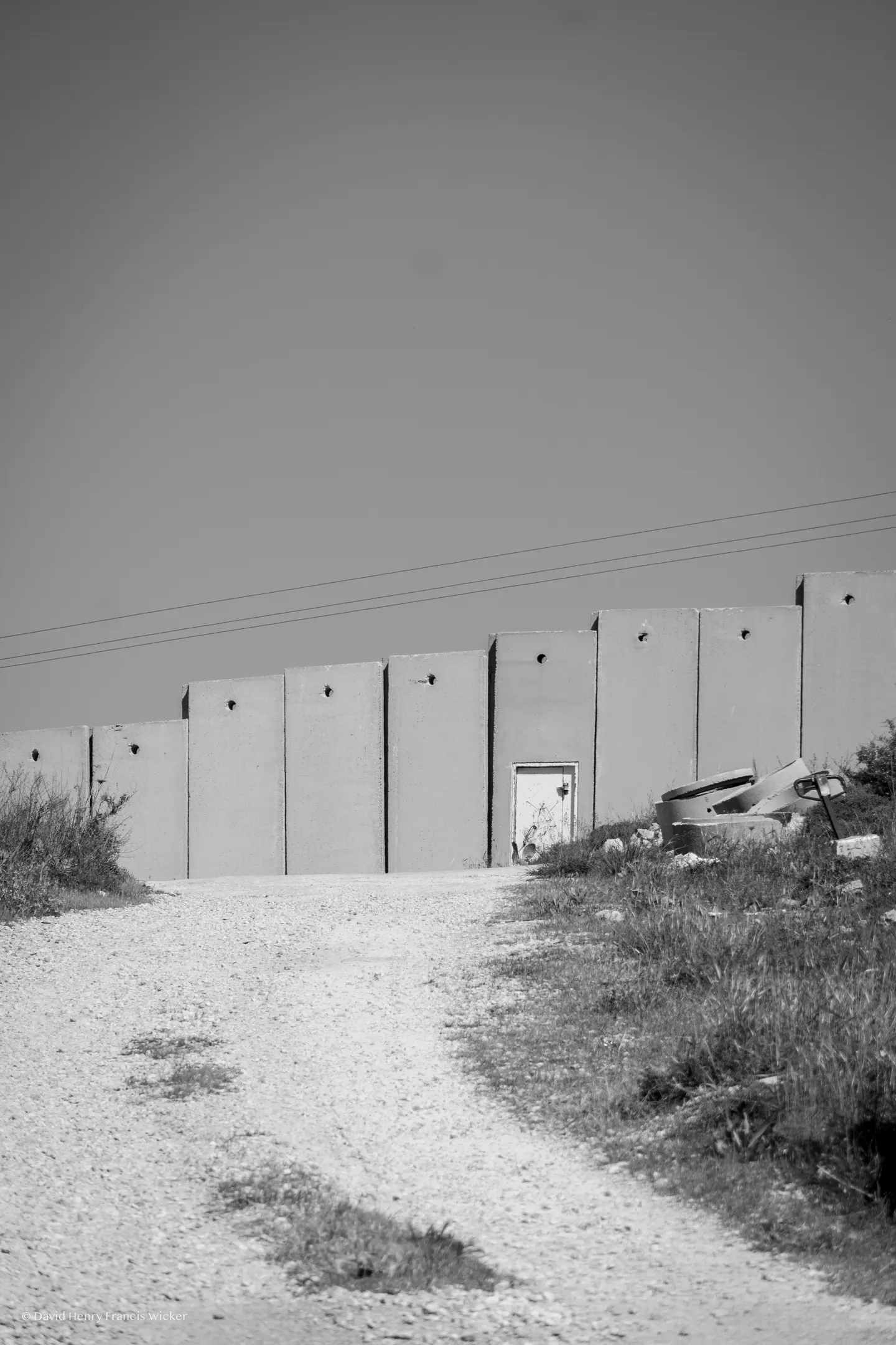 A pale concrete separation wall cuts across a gravel landscape beneath an overcast sky.